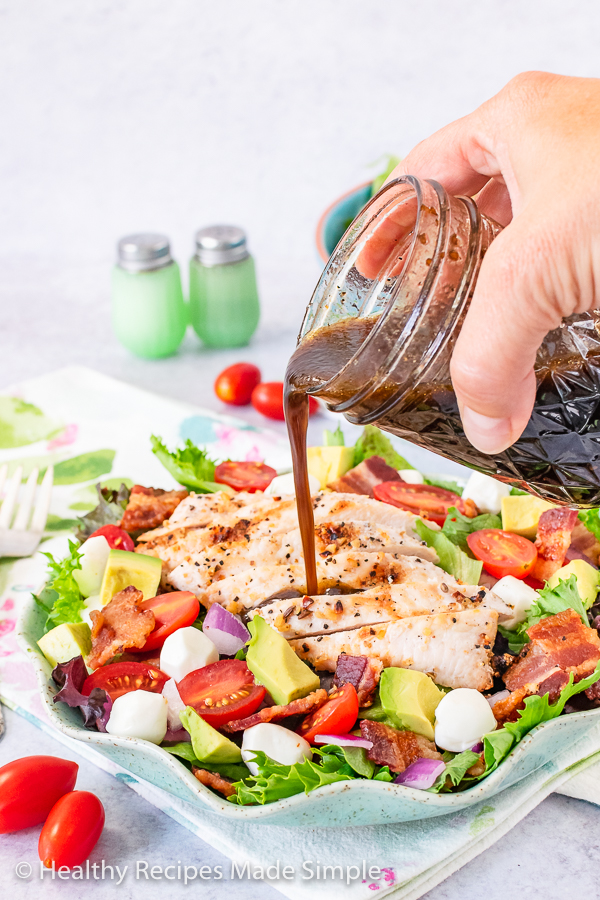 Balsamic dressing being poured from a mason jar onto grilled chicken on a bed of spring mix and vegetables.