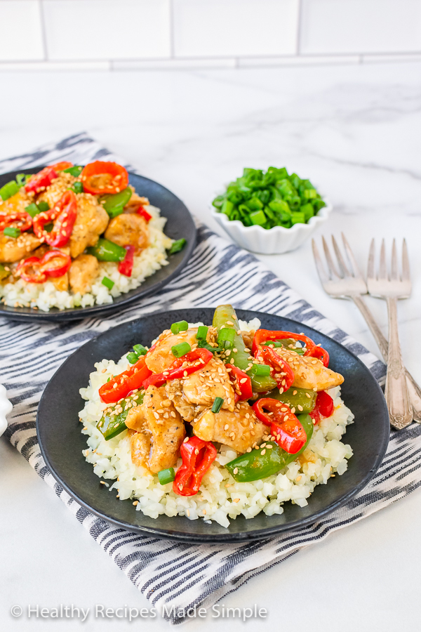 Orange Sesame Chicken on a bed of cauliflower rice served on a black plate with green onions in the background.