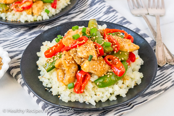 Rectangle image of Orange Chicken on a bed of cauliflower rice served on a black plate with 2 forks.