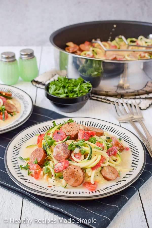 A plate of zucchini noodles with chicken sausage and alfredo sauce with a pan in the background.