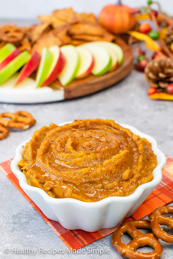 A white bowl full of pumpkin hummus with apples and pretzels in the background.