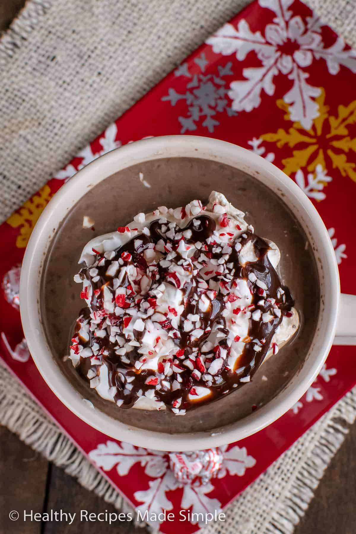 An overhead picture of a cup of hot chocolate topped with whipped cream, chocolate, and peppermint candies.