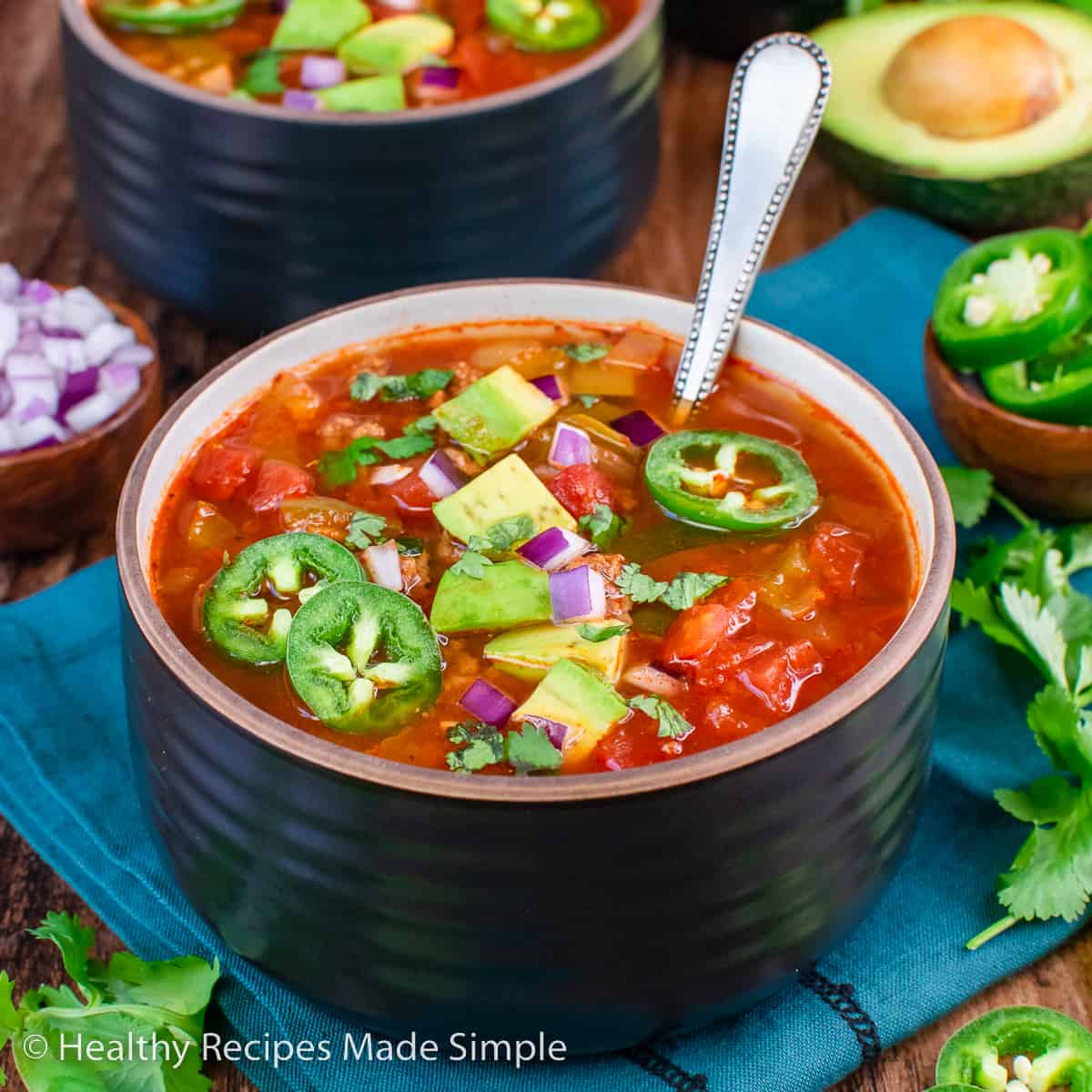 A brown bowl filled with chili without beans topped with diced onions, jalapeños, and avocado.