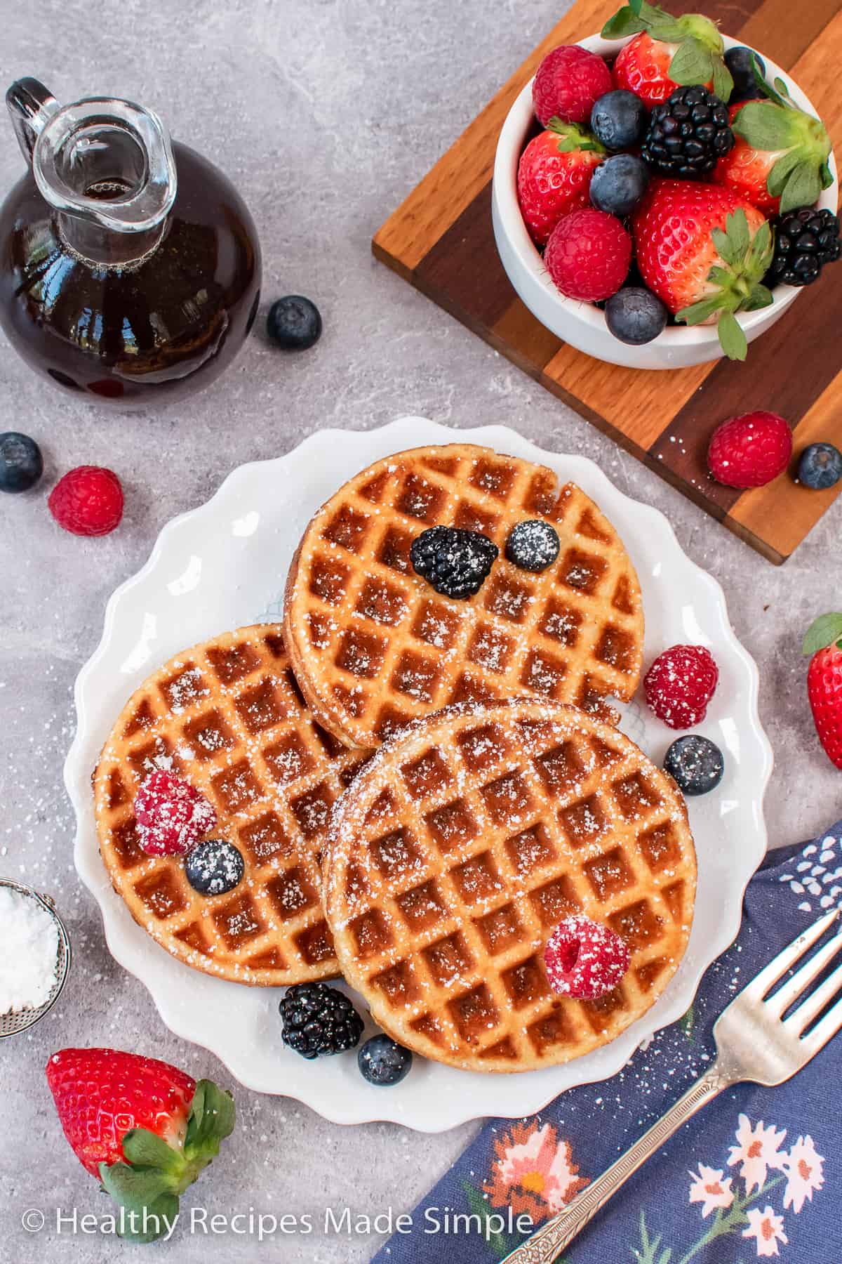 Three waffles on a white plate with powdered sugar and fresh fruit on top.