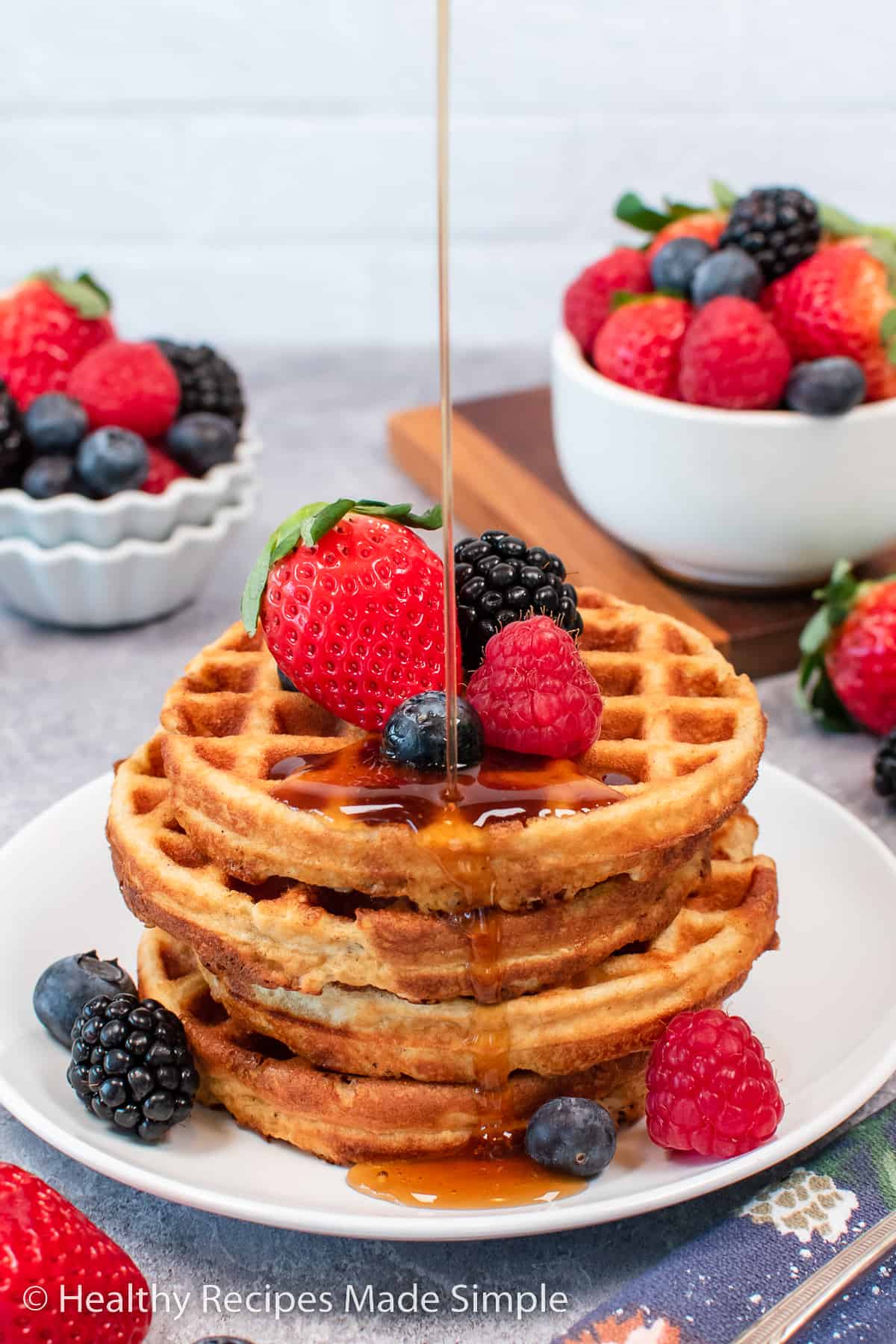 A stack of four waffles on a plate with fresh fruit on top and a stream of syrup.