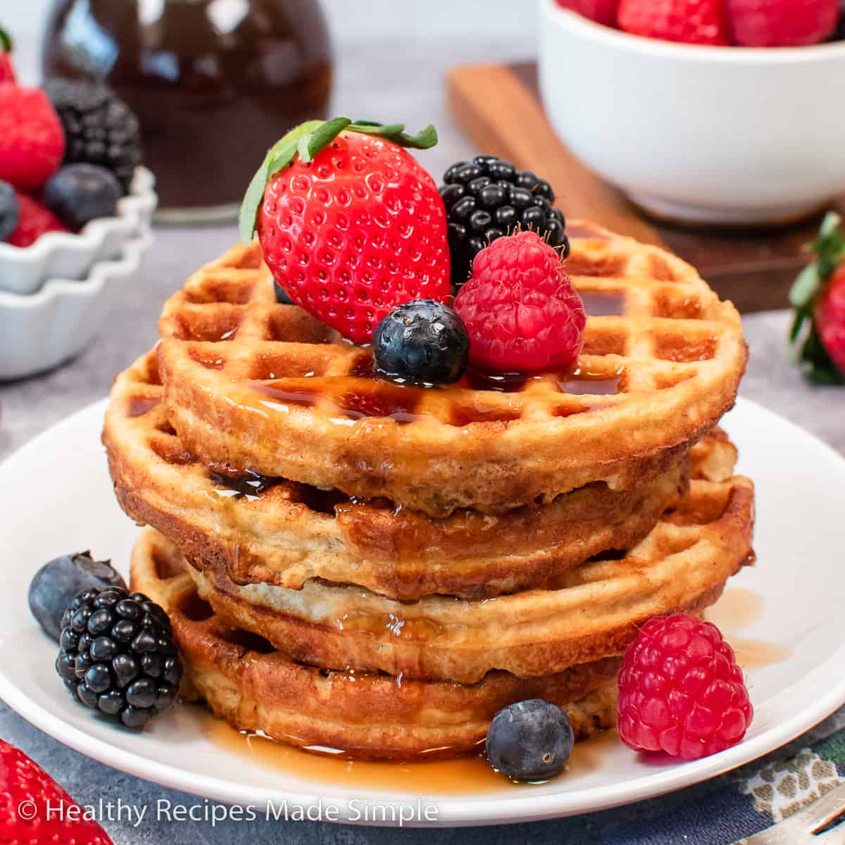 A stack of four waffles on a plate with syrup and fresh fruit on top.