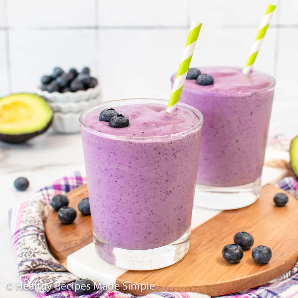 2 glasses of a purple smoothie topped with blueberries and a green and white straw.