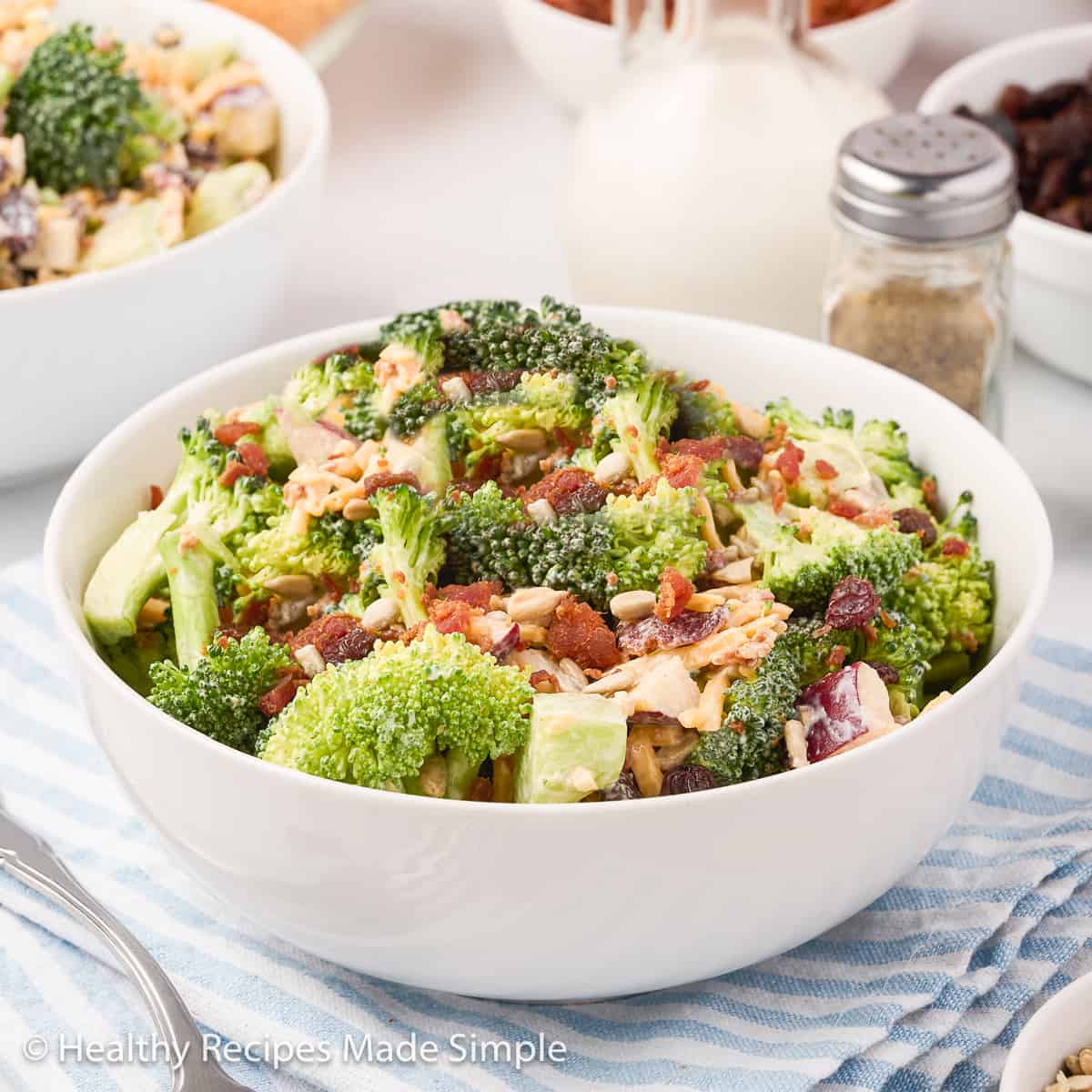 Broccoli salad in a white bowl on a blue and white striped towel.